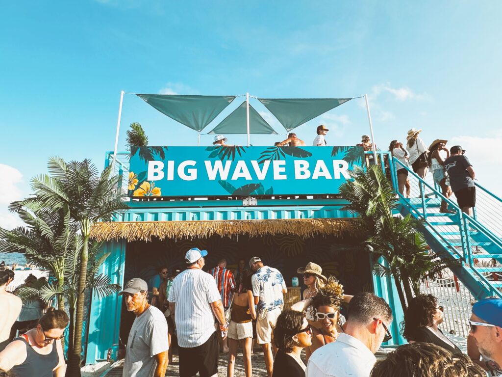 Teal blue Big Wave Bar with people standing on the top patio and a line of guests waiting below.