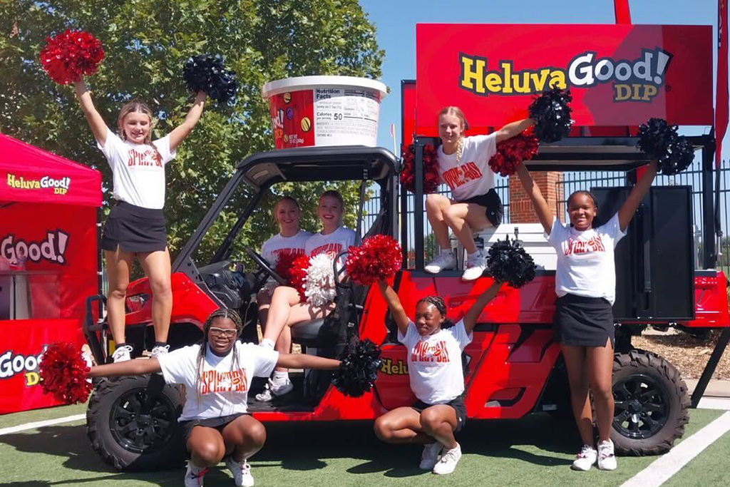 Red Helluva Good Dip promotional cart featuring a large dip container on top, with a group of young cheerleaders posing beside it.
